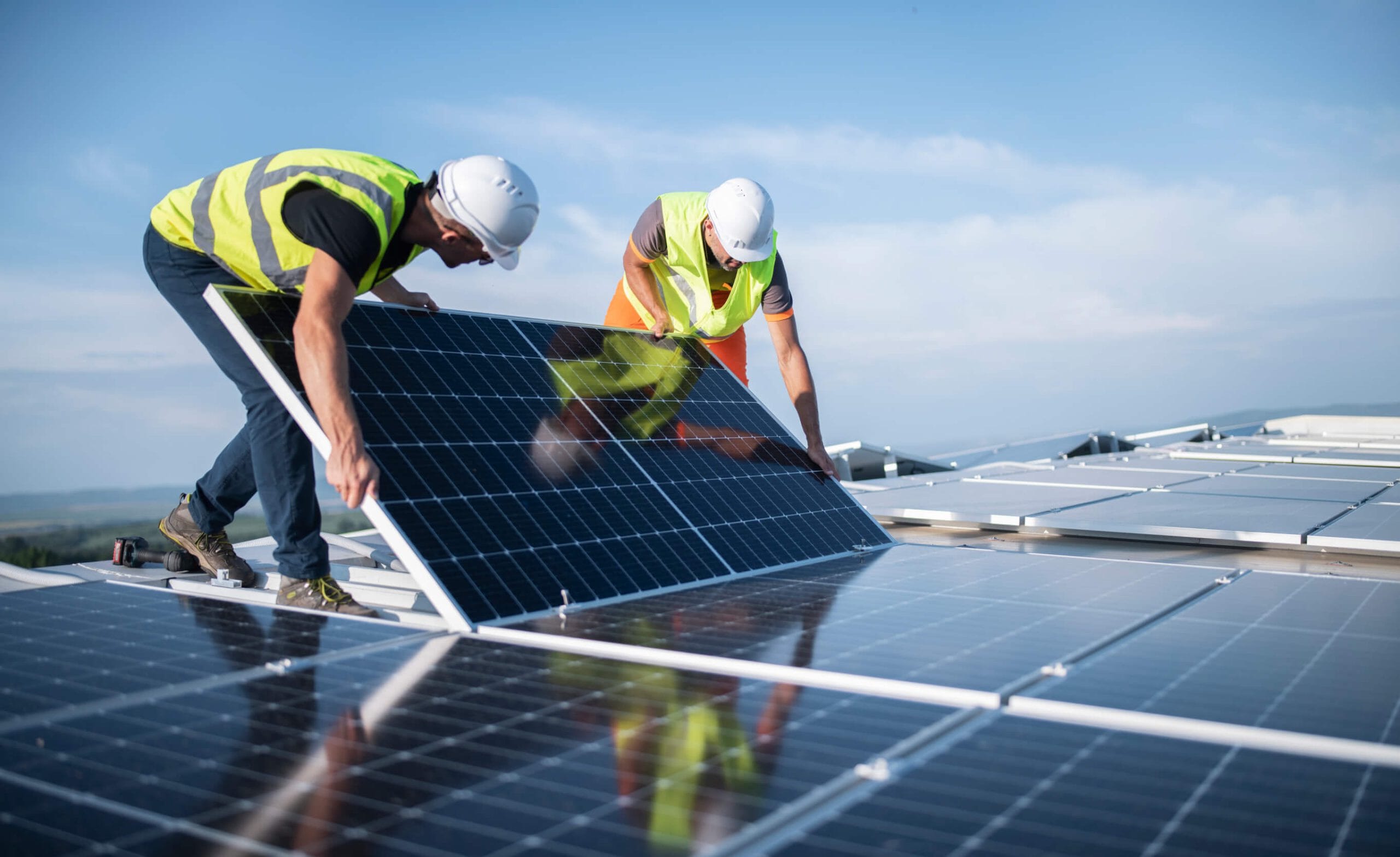 Two engineers in white hard hats fitting a solar panel on a commercial roof, with a blue sky background